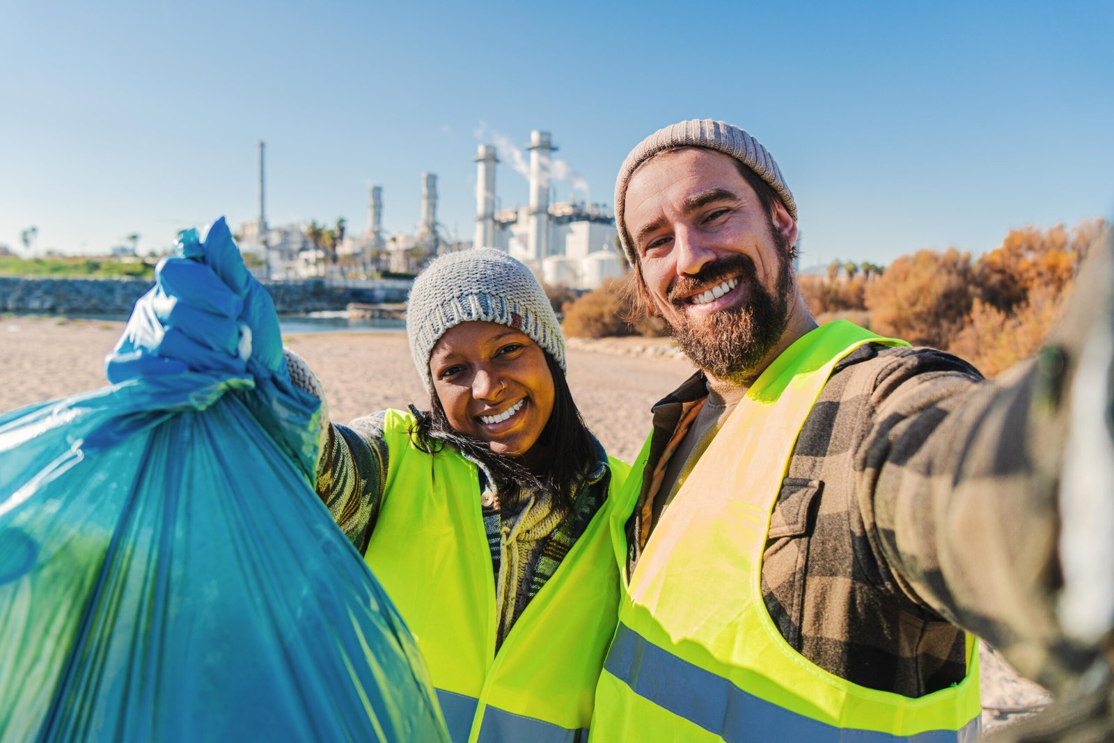 A environmental activist or volunteer multiracial couple picking up trash og the beatch, and taking a selfie portrait smiling looking at camera showing a recycle plastic bag.